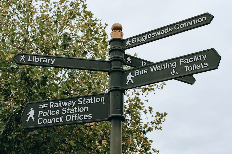 A black metal directional signpost with six signs pointing in various directions is situated outdoors against a background of green tree foliage and a cloudy sky. The signs display information for Biggleswade Common, Library, Railway Station, Police Station, Council Offices, Bus Waiting Facility, and Toilets, with icons indicating pedestrian pathways and accessibility features. The signpost is part of a home relocation or moving logistics setting, as it is adjacent to a street or public area, potentially near a residential or commercial zone used for packing, loading, or transporting furniture and boxes. No vehicles or movers are visible in this image, but the setting suggests an environment where careful planning of moving routes and parking regulations is essential, aligning with services like those provided by Man with Van Gospel Oak for efficient and compliant house removals.