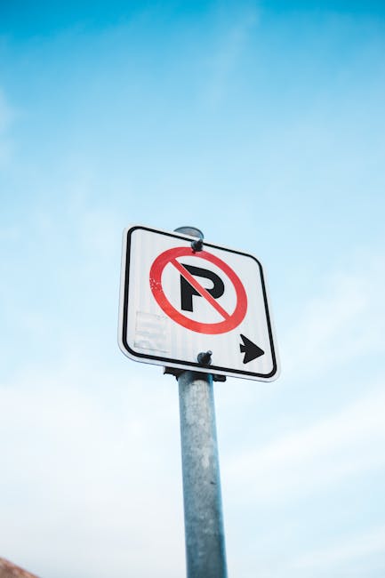 A vertical metal pole supporting a rectangular white traffic sign with a black border, indicating no parking with a red circle and diagonal line crossing out a capital 'P', and a black arrow pointing to the right below. The sign is mounted outside a property, with a clear blue sky in the background. The image is related to house removals and moving logistics, and the sign's presence emphasizes parking restrictions that can impact relocation or furniture transport processes, such as those handled by Man with Van Gospel Oak. The scene highlights the importance of adhering to local parking rules during home relocation or furniture transport to avoid fines, with the sign situated near a residential area that may be relevant for crew parking and loading activities involved in removals services.