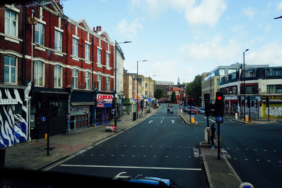 A street scene on Gospel Oak Road in London, showing a mix of commercial buildings and residential apartments. On the left side, there are shopfronts with colorful signage, some with black and white striped designs, and one with a red and white 'Carolina' sign. The buildings are brick with multiple windows, and there are streetlights above. The street has a dedicated lane marked for vehicles, with traffic signals displaying red lights at the pedestrian crossing. The pavement on both sides has pedestrians, some waiting at the crossing or walking along the sidewalk. Vehicles are visible in the distance, driving and waiting at the lights. The environment is illuminated by natural daylight with a partly cloudy sky overhead. This scene represents an urban setting suitable for home relocation or furniture transport activities, with visible loading areas and surrounding infrastructure that Man with Van Gospel Oak might utilize during moving and packing processes.