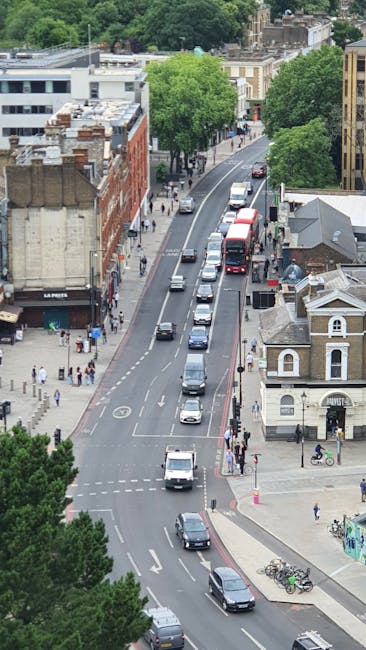 An aerial view of Gospel Oak Road in London showing a mixture of residential and commercial buildings, including a brick building with several stories on the left and smaller houses on the right. The street has a single lane of traffic moving in each direction, with cars, vans, and a double-decker bus visible, along with parked vehicles along the curb. Pedestrians are walking on the sidewalks, some crossing the street at designated crosswalks. Green trees line both sides of the road, providing shade and contrast to the urban setting. The image captures the busy scene during daylight, with natural lighting illuminating the buildings, vehicles, and pedestrians, relevant to the process of home relocation or furniture transport by companies such as Man with Van Gospel Oak during packing and loading activities on Gospel Oak Road.