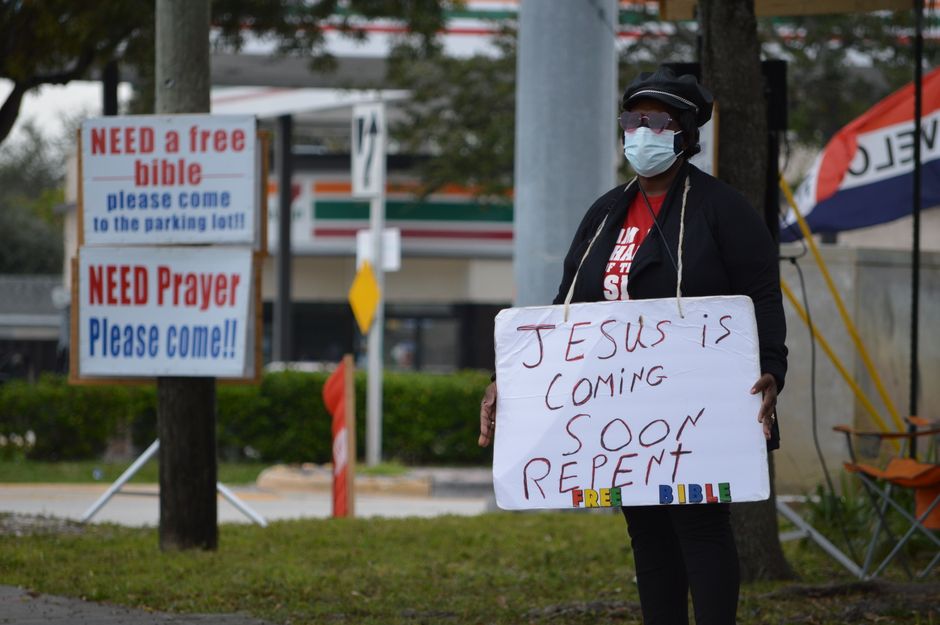A person wearing a face mask, black jacket, and sunglasses is standing outdoors on a sidewalk, holding a large white cardboard sign with the handwritten message 'JESUS IS COMING SOON REPENT.' In the background, there are two informational signs: one reading 'NEED a free bible please come to the parking lot' and another reading 'NEED Prayer Please come!!,' both attached to a wooden post. The environment includes a grassy area, trees, a street with visible traffic lights, and a 7-Eleven store, suggesting a suburban setting. The individual appears to be participating in a public religious outreach or street preaching, with a focus on religious messages and community engagement, in a scene related to house removals and moving logistics contextually aligned with the idea of preparation and planning.