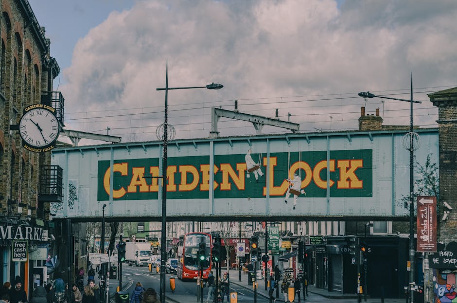 A large, weathered sign reading 'Camden Lock' in bold orange capital letters on a blue-green metal bridge spans across a busy street in Camden, London, with pedestrians walking below. The bridge is supported by metal beams and has two street lamps attached, along with two hanging streetlights overhead. To the left, a classic street clock displays the time as approximately 11:50 and is mounted on a building with traditional brickwork and balcony railings. Below the bridge, various shops, signs, and adverts are visible, including a cash point and a 'Market' sign. There are numerous pedestrians on the pavement, some waiting at traffic lights, while others walk along the street. A red double-decker bus and other vehicles are present, with the overall scene reflecting a lively urban atmosphere. The overcast sky is filled with grey clouds, creating diffused lighting over the area, which is typical of a busy London street setting, relevant to furniture transport and home relocation logistics. Man with Van Gospel Oak may assist with such moving processes as indicated by the surroundings suggesting busy street scene and transport activity.