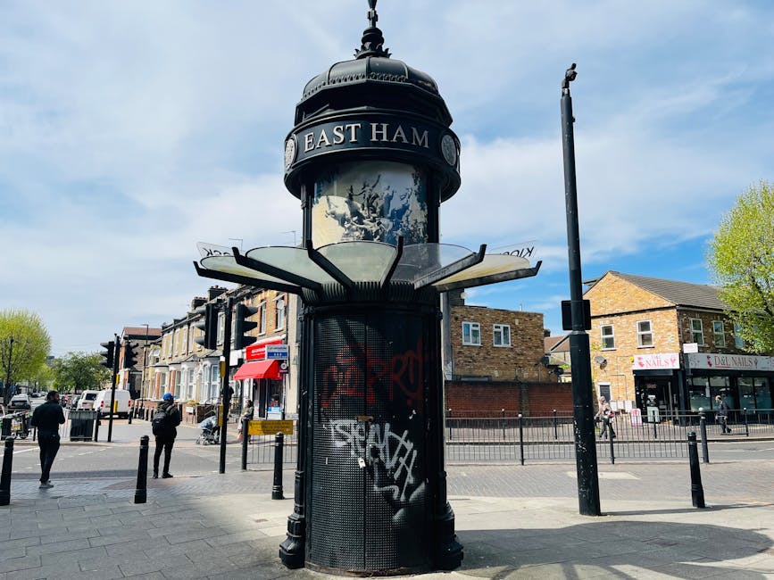A historic black cylindrical tram stop shelter with a domed top, displaying a sign reading 'EAST HAM' in white letters, situated on a busy urban street in Gospel Oak. The shelter has a decorative design with a mesh exterior and is covered in colorful graffiti tags. Inside, several cardboard boxes and plastic-wrapped furniture or goods can be seen, likely part of ongoing home relocation or packing and moving activities. To the right of the shelter, a black lamppost and street signs are visible, with pedestrians walking on the pavement nearby. In the background, there are residential buildings, small commercial shops, and trees with fresh spring foliage under a partly cloudy sky. Man with Van Gospel Oak occasionally uses such locations as part of its furniture transport and loadings during house removals or moving services, supporting efficient packing and moving logistics.