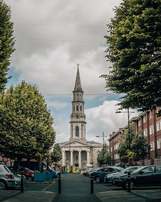 A street view in Gospel Oak featuring a church with a tall, pointed spire and classical architectural details, positioned centrally in the background. The street is lined with red-brick residential buildings on the right side and mature green trees on both sides, creating a canopy over the pavement. Several parked cars are visible along the sides of the street, and the foreground shows a loading area with a blue plastic container, cardboard boxes, and blankets used for protection during a home relocation. There are no moving personnel or equipment in the image, but the scene suggests a preparatory phase of furniture transport or packing and moving processes, possibly associated with a professional removals service like Man with Van Gospel Oak for house removal or last-minute move arrangements. The sky above is partly cloudy, with natural daylight illuminating the scene, emphasizing the urban environment and the logistics involved in house removals within the area.