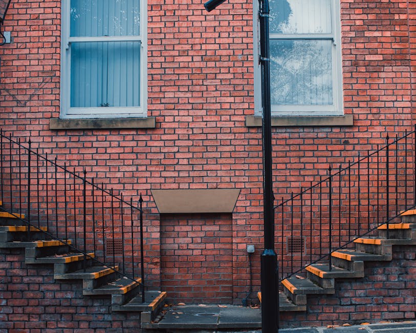 Exterior view of a red brick residential building featuring two windows with white frames and window sills, a central rectangular blind or cover on the brick wall, and two external staircase fire escapes with metal stairs and black railings on either side, leading to an upper doorless platform. The stairs have dark treads with yellow safety strips along the edges, and the surrounding environment includes a stone or concrete pavement with some fallen leaves. The scene is evenly lit with natural daylight, and the staircase setup is typical of urban home access. The image captures the outdoor environment at ground level, relevant to house removal preparations or building access during a home relocation process, reflecting the challenges of tight staircases between floors that professional movers like Man with Van Gospel Oak often manage during furniture transport and packing and moving services.