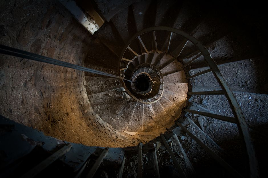 A view looking down a narrow, winding staircase within a residential building featuring old, worn brick walls and a metal handrail attached to the wall on the right side. The staircase has a spiral shape, with steps made of wood or concrete, leading downwards into darkness. The environment appears dimly lit, with natural light or a small light source illuminating the top portion of the stairs. This internal staircase is part of a home undergoing a moving process, with the image capturing the challenging, tight space that Man with Van Gospel Oak may need to navigate during a house removal or furniture transport. The setting highlights the importance of careful planning and equipment use in moving furniture through confined interior staircases during home relocations or packing and moving tasks.
