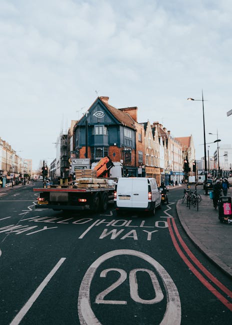 A white flatbed truck with a black chassis is parked on the street in Gospel Oak, containing wooden pallets and large cardboard boxes secured with plastic wrap for a home relocation. The truck is positioned at an intersection with clearly marked road signs, including a prominent 20 mph speed limit circle painted on the road surface. Nearby, a white van is parked adjacent to the truck, with its rear doors closed. The surrounding environment includes a row of mixed-use buildings with brick facades and shops at ground level, some with awnings and signage, and residential windows above. Several pedestrians are walking along the pavement, and bicycles are parked nearby. The open urban street scene is illuminated by natural daylight, and outdoor streetlights are visible. This scene captures part of the packing and loading process involved in furniture transport and house removals, with the presence of equipment such as trolleys or straps not visible in this image. The mention of Man with Van Gospel Oak is relevant in the context of professional removals services supporting house moves in NW5.