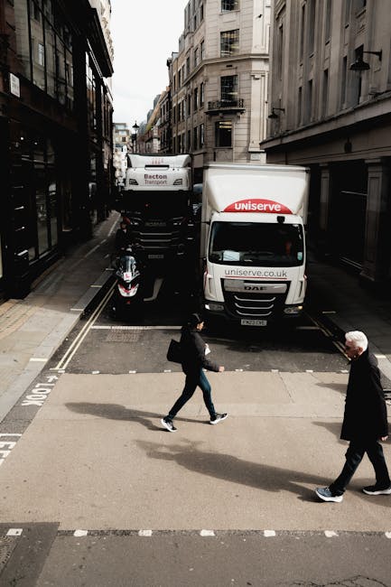 A street scene showing a white delivery van marked with 'uniserve.co.uk' parked partially on the pavement and road, with its rear doors closed. Behind it, a larger vehicle with signs indicating it belongs to Becton Transport is visible. The vehicles are positioned near a narrow urban street lined with multi-storey buildings on both sides, featuring fire escapes and large windows. In the foreground, two pedestrians are crossing the street: a woman wearing a black jacket and jeans carrying a bag, and a man dressed in a dark suit with grey hair. The scene occurs during daytime with natural light illuminating the environment, which may relate to home relocation or furniture transport activities managed by Man with Van Gospel Oak, especially involving the loading or unloading process of house removals in the NW5 area.